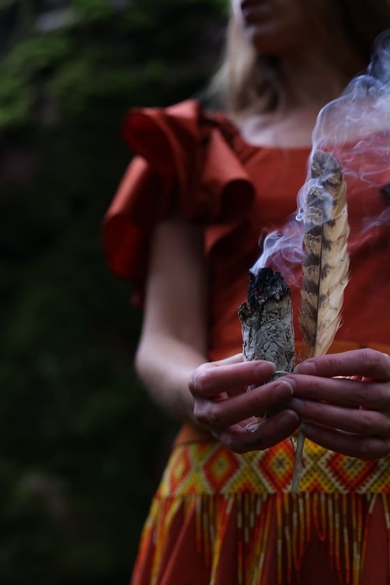Close-up of hands holding a smoking herb bundle and a striped feather above a colourful beaded belt.